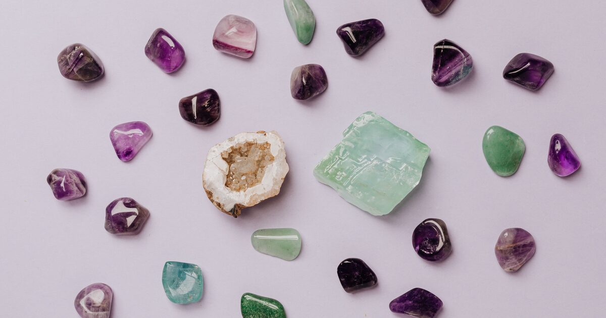 An assortment of tumbled amethyst, aventurine, and quartz gemstones with a geode on a lilac surface