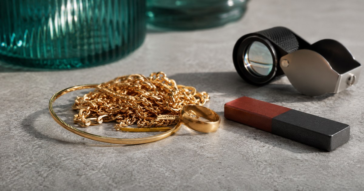 A heap of gold-colored chains, a bangle, and a plain band on a stone surface next to a bar magnet and a loupe