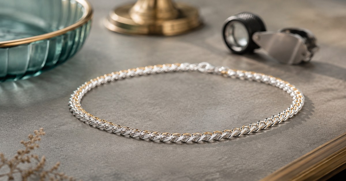 A sterling silver chain necklace with a warm gold sheen along its edges, resting on a stone surface beside a jeweler's loupe